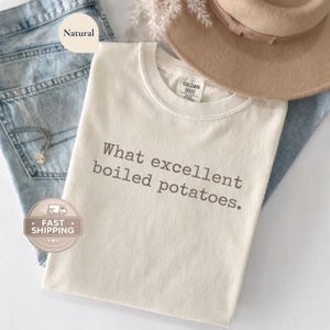 Puede incluir: Camiseta color crema con el texto "What excellent boiled potatoes". La camiseta está doblada sobre unos vaqueros azules. También se ven un sombrero beige y un collar plateado. La camiseta es de un material suave.