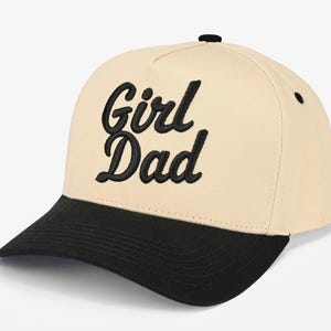 May include: A beige baseball cap with a black brim and a black button on top. The front of the cap features the text Girl Dad embroidered in black script lettering. The cap has a structured crown and a curved visor, providing a classic look.