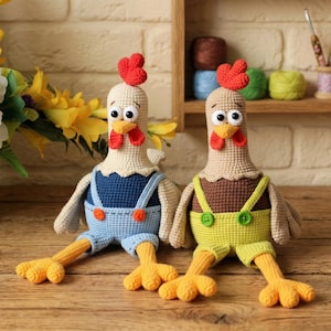 Op de afbeelding: Twee handgemaakte gehaakte kippenpoppen met grote ogen, rode kammen en gele voeten. De ene kip draagt een blauwe overall, de andere een gele overall. De kippen zitten op een houten oppervlak.