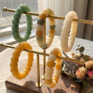 Könnte beinhalten: Ein Displayständer mit fünf Armbändern aus grünen, gelben und weißen Perlen. Die Armbänder bestehen aus abgerundeten Perlen. Der Ständer ist aus goldfarbenem Metall auf einem Holzsockel. Ein Blumenstrauß ist im Hintergrund.