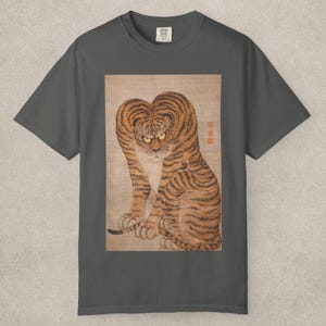 May include: Dark grey t-shirt featuring a vintage illustration of a tiger. The tiger is depicted with orange and black stripes, yellow eyes, and a stern expression. The artwork is printed on a rectangular patch.
