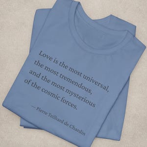 May include: A light blue t-shirt with the quote "Love is the most universal, the most tremendous, and the most mysterious of the cosmic forces." by Pierre Teilhard de Chardin printed in black.