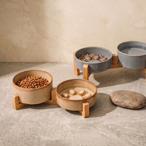 May include: Two elevated pet feeding stations, each featuring two ceramic bowls held by a light-colored wooden stand. One station has beige bowls, one filled with dry kibble and one with a paw-shaped slow feeder. The other has grey bowls.