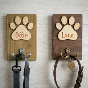 May include: Two wooden pet leash holders. One is stained a light brown with a paw print and the name "Ollie". The other is stained a darker brown with a paw print and the name "Luna". Each has a metal hook.