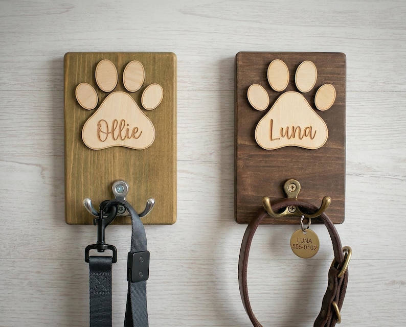 May include: Two wooden pet leash holders. One is stained a light brown with a paw print and the name "Ollie". The other is stained a darker brown with a paw print and the name "Luna". Each has a metal hook.