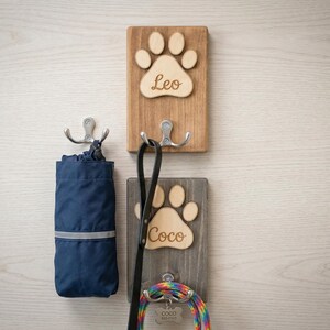 May include: Wooden wall-mounted dog leash and accessory holder. The holder features two wooden plaques with paw print cutouts and names "Leo" and "Coco". A blue bag, a black leash, and a colorful leash are hanging from the hooks.