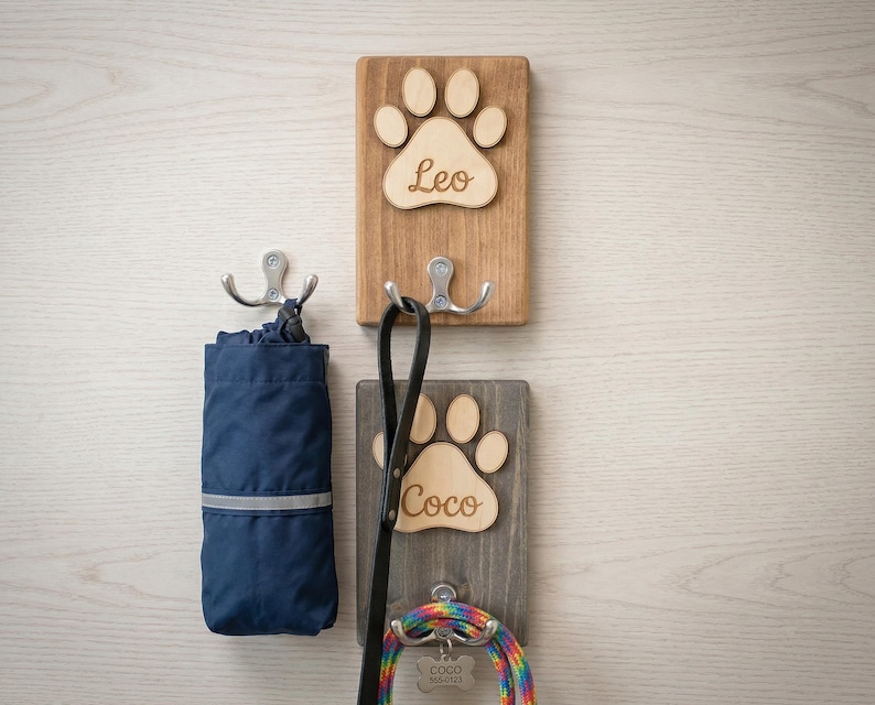 May include: Wooden wall-mounted dog leash and accessory holder. The holder features two wooden plaques with paw print cutouts and names "Leo" and "Coco". A blue bag, a black leash, and a colorful leash are hanging from the hooks.