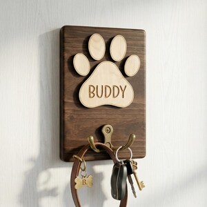 May include: A wooden wall-mounted dog leash holder with a paw print design and the name "BUDDY". The holder has two hooks for leashes and keys, and a small bone-shaped tag. The wood is dark brown with a light brown paw print.