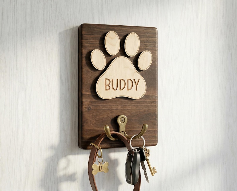 May include: A wooden wall-mounted dog leash holder with a paw print design and the name "BUDDY". The holder has two hooks for leashes and keys, and a small bone-shaped tag. The wood is dark brown with a light brown paw print.