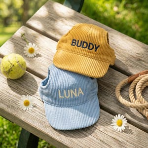 May include: Two corduroy dog hats, one mustard yellow with "BUDDY" embroidered, and one light blue with "LUNA" embroidered. A tennis ball, daisies, and a rope leash rest on a wooden bench.