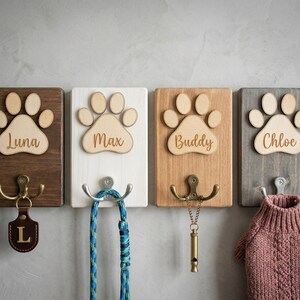 May include: Four wooden pet leash holders with paw print designs. Each holder has a different color and name: Luna, Max, Buddy, and Chloe. Each has a different hook and accessory, including a leash, whistle, and dog sweater.