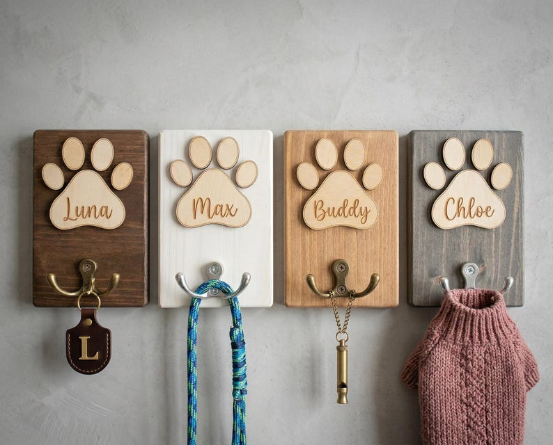May include: Four wooden pet leash holders with paw print designs. Each holder has a different color and name: Luna, Max, Buddy, and Chloe. Each has a different hook and accessory, including a leash, whistle, and dog sweater.