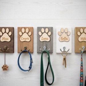 May include: Five wooden pet leash holders with paw print designs and names: Max, Cooper, Luna, Bella, and Milo. Each holder has a metal hook and is mounted on a white wooden wall. Accessories include a leash, collar, and keys.