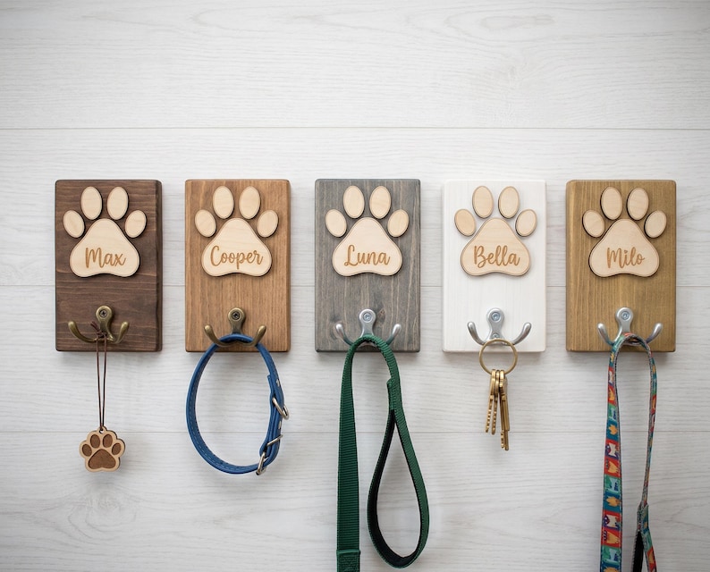 May include: Five wooden pet leash holders with paw print designs and names: Max, Cooper, Luna, Bella, and Milo. Each holder has a metal hook and is mounted on a white wooden wall. Accessories include a leash, collar, and keys.