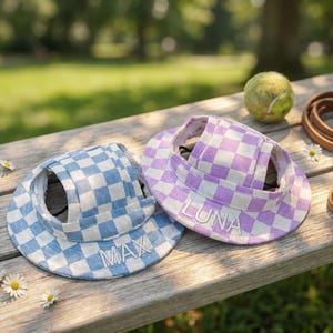 May include: Two dog visors with a checkered pattern in blue and purple, each with a name embroidered on the brim. A tennis ball and a leather leash are also on the wooden surface. Daisies are scattered around.