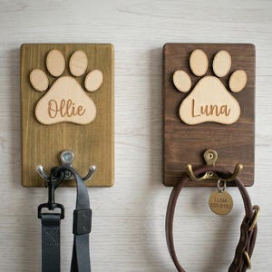 May include: Two wooden pet leash holders. One is stained a light brown with a paw print and the name "Ollie". The other is stained a darker brown with a paw print and the name "Luna". Each has a metal hook.