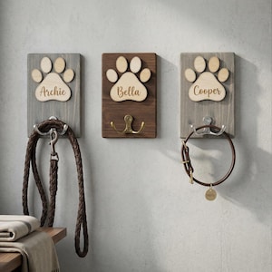 Pode incluir: Três suportes de madeira para trelas e coleiras de animais de estimação. Cada um tem um design de pegada e o nome de um animal de estimação: Archie, Bella e Cooper. Uma trela de couro castanho pende do suporte de Archie. O suporte de Bella tem um gancho de latão. O suporte de Cooper tem uma coleira castanha.