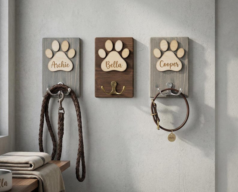 May include: Three wooden pet leash and collar holders. Each has a paw print design and a pet's name: Archie, Bella, and Cooper. A brown leather leash hangs from Archie's holder. Bella's holder has a brass hook. Cooper's holder has a brown collar.