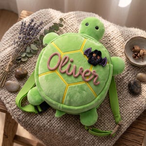 May include: A plush green turtle-shaped backpack featuring a soft shell design with yellow piping. The name Oliver is embroidered in a textured pink script across the center, accompanied by a small black bat patch with purple wings.
