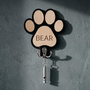 May include: Wooden paw print key holder with the word "BEAR" engraved on the centre pad. The paw print is black with light wood-coloured pads. It has two hooks and a silver whistle attached.