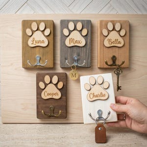 May include: A wooden wall-mounted key holder with five individual key hooks. Each hook has a paw print design with a name. The names are Luna, Max, Bella, Cooper, and Charlie. The key holder is made of wood and has a natural finish.
