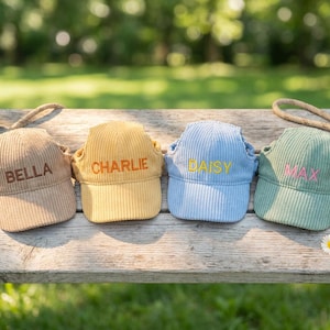 May include: Four corduroy dog hats in beige, yellow, light blue, and green, each embroidered with a different name: Bella, Charlie, Daisy, and Max. A tennis ball and leashes are also visible.