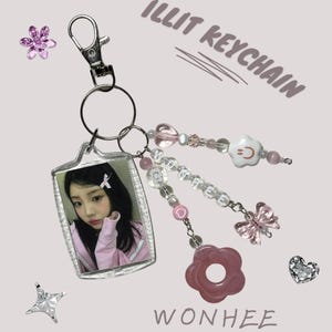 May include: A clear acrylic keychain with a photo of a person in a pink top. The keychain has a silver clasp and is adorned with pink and clear beads, a pink flower, and a white flower with a smiley face. The text "ILLIT KEYCHAIN" and "WONHEE" are visible.