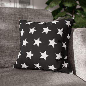 May include: A square throw pillow featuring a solid black background with a repeating pattern of white five-pointed stars. The pillow has a soft, plush appearance and is positioned upright, displaying a consistent star print across the front surface.