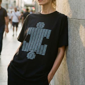 May include: Black t-shirt featuring a white graphic made of repeated four-letter acronyms. The model is leaning against a grey wall. The background shows a street scene with blurred figures and buildings.