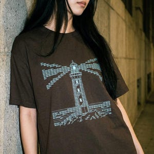 May include: A brown t-shirt with a lighthouse design. The graphic is in light blue and features a minimalist lighthouse with text. The shirt has a crew neck and short sleeves.