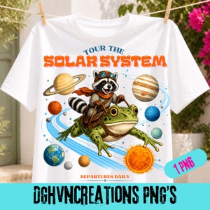 Peut inclure: T-shirt blanc avec un motif fantaisiste d'un raton laveur portant des lunettes et une écharpe, chevauchant une grenouille à travers le système solaire. Le texte "Tour the Solar System" est en orange, avec des planètes et des étoiles. Le bas du t-shirt porte l'inscription "Departures Daily".
