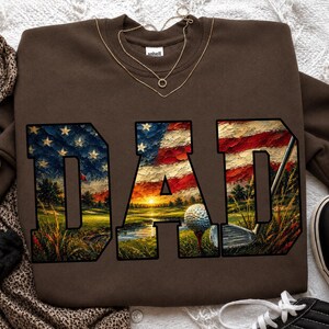 Peut inclure: Un sweat-shirt marron avec le mot "DAD" en grandes lettres. Les lettres sont remplies d'une image du drapeau américain et d'une scène de terrain de golf, avec une balle de golf, un club et un coucher de soleil.