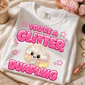 Puede incluir: Una camiseta blanca con un estampado gráfico que incluye el texto "YOU'RE A GLITTER DUMPLING" en letras rosas gruesas. El diseño presenta una ilustración central de una bola de masa redonda, iridiscente y brillante con cara sonriente y estrellas rosas.