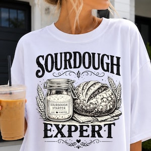 Op de afbeelding: Wit T-shirt met een grafisch ontwerp met het woord "SOURDOUGH" boven een illustratie van een pot zuurdesemstarter en een brood. Het woord "EXPERT" staat onder de illustratie.
