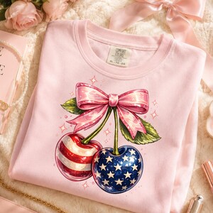 Peut inclure: Un sweat-shirt rose clair Comfort Colors avec un motif de deux cerises et un nœud rose. Une cerise a des rayures rouges et blanches, l'autre a des étoiles bleues et blanches. Le sweat-shirt a un col rond.