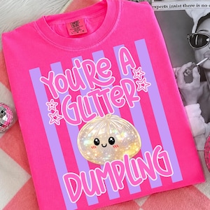 以下が含まれることがあります： 明るいピンク色のTシャツで、縦の薄紫色のストライプが入ったグラフィックプリントが施されています。ピンク色の文字で「YOU'RE A GLITTER DUMPLING」と書かれており、小さな星のアイコンと、笑顔で輝くダンプリングのイラストが中央に描かれています。