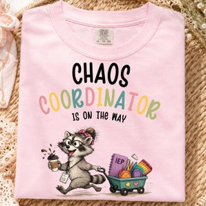 Peut inclure: Un t-shirt rose clair avec une illustration de raton laveur portant un nœud rose et des lunettes, tenant un café. Le texte "CHAOS COORDINATOR IS ON THE WAY" est imprimé au-dessus du raton laveur qui tire un chariot rempli de fournitures.