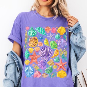 Peut inclure: Un t-shirt violet orné d'un motif de coquillages colorés. Le graphisme comprend divers coquillages, étoiles de mer et algues dans des tons de jaune, orange, vert, bleu et violet. Le t-shirt a des manches courtes et une coupe ample.