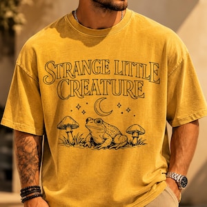 Peut inclure: T-shirt jaune moutarde avec l'inscription "Strange Little Creature". Le motif comprend une grenouille, des champignons, un croissant de lune et des étoiles. Le t-shirt a des manches courtes et une coupe décontractée.