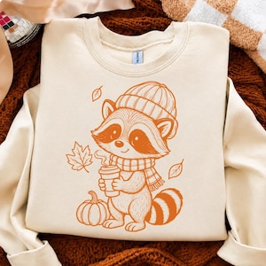 Peut inclure: Un sweat-shirt à col rond couleur crème arborant un motif orange brûlé représentant un raton laveur portant un bonnet en tricot et une écharpe. Le raton laveur tient une tasse de café fumante près d'une citrouille et de feuilles d'automne.