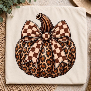 Peut inclure: Un t-shirt couleur crème présentant un motif central de citrouille avec un imprimé léopard. La citrouille est ornée d'un grand nœud proéminent avec un motif à carreaux marron et beige, centré sur le haut de la citrouille.