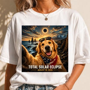 May include: A white t-shirt featuring a graphic of a golden retriever wearing sunglasses and an American flag bandana, taking a selfie against a coastal landscape during a total solar eclipse, with the text TOTAL SOLAR ECLIPSE AUGUST 12, 2026.