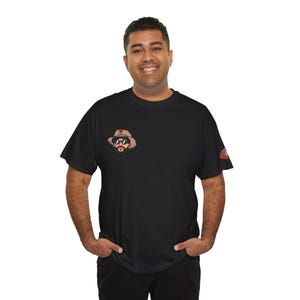Peut inclure: T-shirt noir avec une illustration de dessin animé d'un homme portant un chapeau et des lunettes de soleil sur la poitrine gauche. Un petit motif est sur la manche droite.
