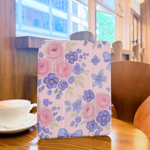 May include: A light pink tablet case adorned with a watercolor floral pattern. The design features large pink roses and clusters of blue flowers with green leaves. The case is on a wooden table, with a cup of coffee in the foreground.