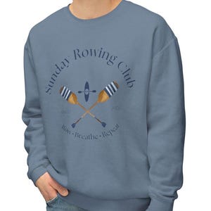 Puede incluir: Sudadera azul con el texto "Sunday Rowing Club" en un diseño circular. El diseño incluye remos cruzados, un kayak y las palabras "Row. Breathe. Repeat."