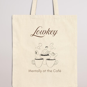 Könnte beinhalten: Cremefarbene Tragetasche mit dem Wort "Lowkey" in brauner Schrift. Darunter eine Grafik, die Menschen zeigt, die sich in Kaffeetassen entspannen, mit dem Text "Mentally at the Café". Die Tasche hat zwei Griffe.