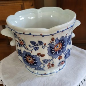 Może przedstawiać: Biała ceramiczna doniczka z falistym brzegiem i dwoma uchwytami po bokach, ozdobiona powtarzającym się wzorem niebieskich i brązowych motywów kwiatowych z liściastymi pnączami. Naczynie ma zaokrąglony, żłobkowany korpus oraz gładkie, błyszczące wykończenie.