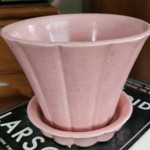 May include: A light pink ceramic flower pot with a matching saucer. The pot has a fluted design and a speckled finish. The saucer has a scalloped edge. The pot is designed to hold plants and flowers.