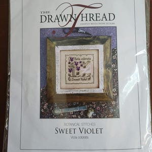 May include: A needlework pattern packet titled The Drawn Thread Sweet Violet Viola odorata. The cover features a cross-stitch design of purple flowers in a wooden frame, with a small metal thimble and measuring tool visible below the frame.