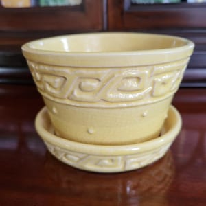 Könnte beinhalten: Ein gelber Keramik-Blumentopf mit passender Untertasse. Der Topf hat ein dekoratives geometrisches Muster am oberen und unteren Rand mit kleinen erhabenen Punkten. Das Set ist aus Keramik und für den Innen- und Außenbereich geeignet.